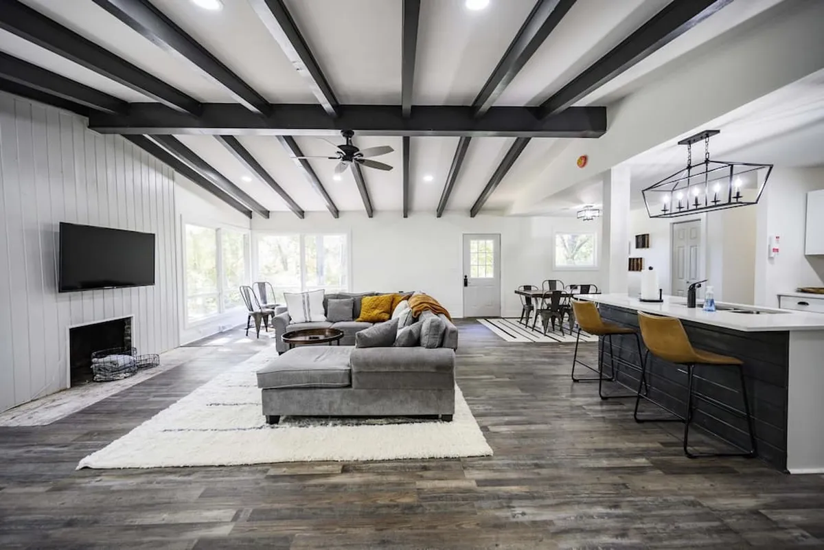 The hero image showcases a spacious, sunlit living area with a large, plush sectional sofa arranged for conversation, a modern dining table ideal for shared meals, beautiful exposed wooden beams overhead, and a sleek wall-mounted television. The room exudes warmth and is designed for togetherness, perfect for relaxing, socializing, or gathering with family and friends.