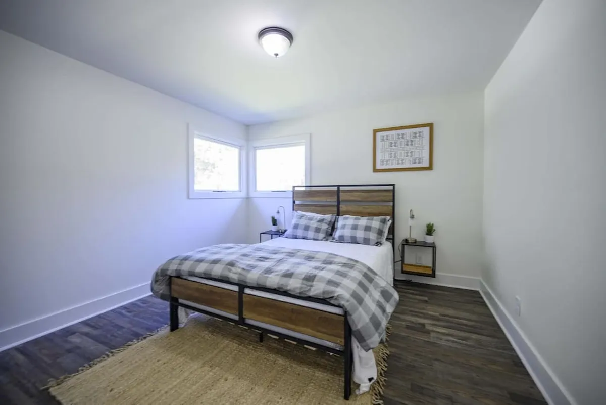 A tranquil bedroom scene with a bed dressed in plaid bedding, a nightstand and lamp, framed wall art, and windows that let in warm natural light. The atmosphere is cozy and peaceful, ideal for relaxation or quiet moments of reading.