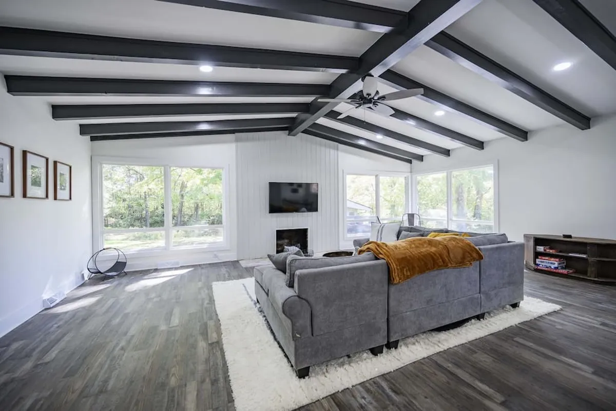 The photo features a spacious living room with a plush sofa, a wall-mounted television, expansive windows, and prominent ceiling beams. The space exudes warmth and comfort, inviting group relaxation or casual conversation.