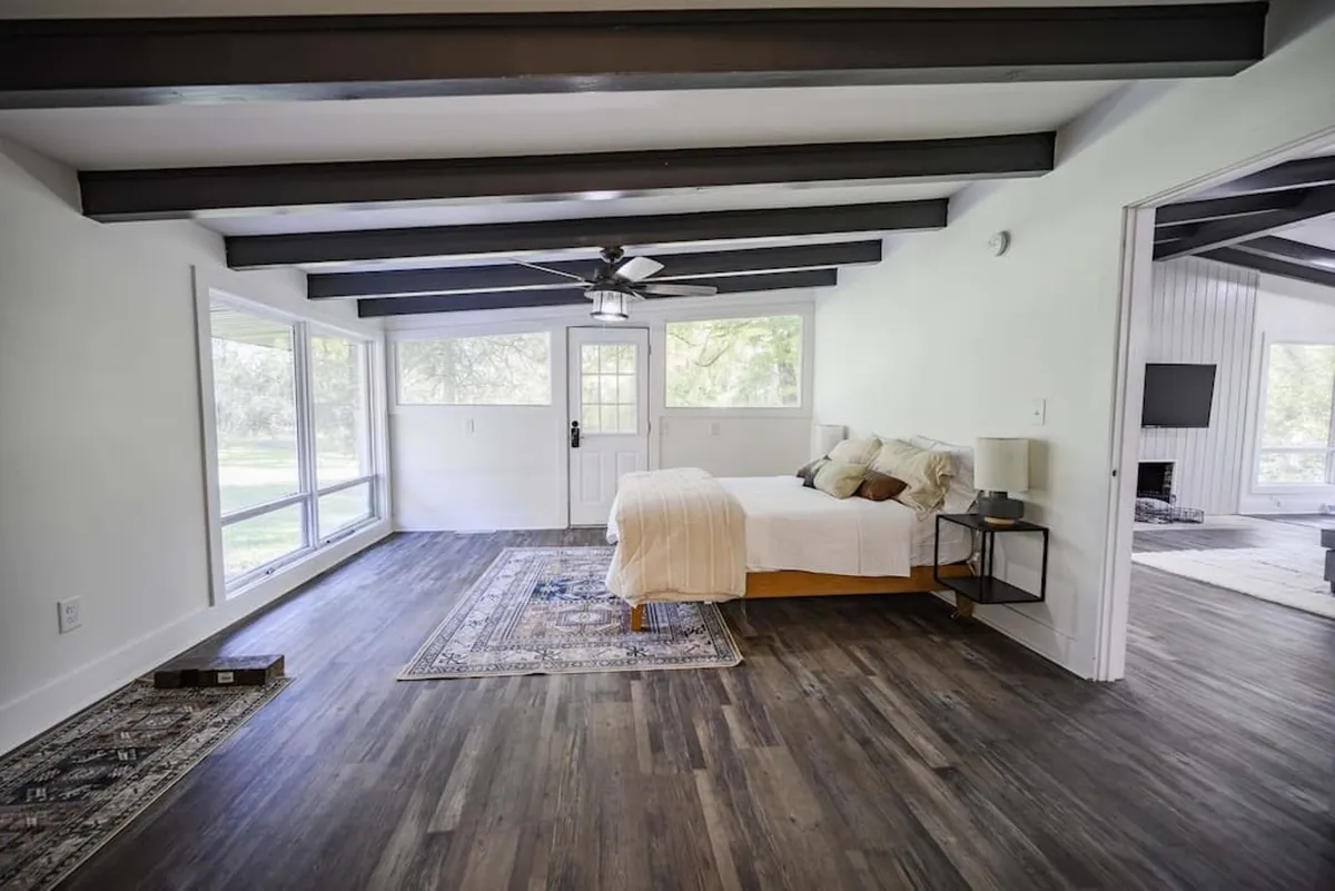 A serene bedroom featuring a bed near large windows that invite in natural light and offer views of the surrounding trees. The space is accented by exposed ceiling beams and a soft rug, creating a peaceful retreat that feels connected to the outdoors.