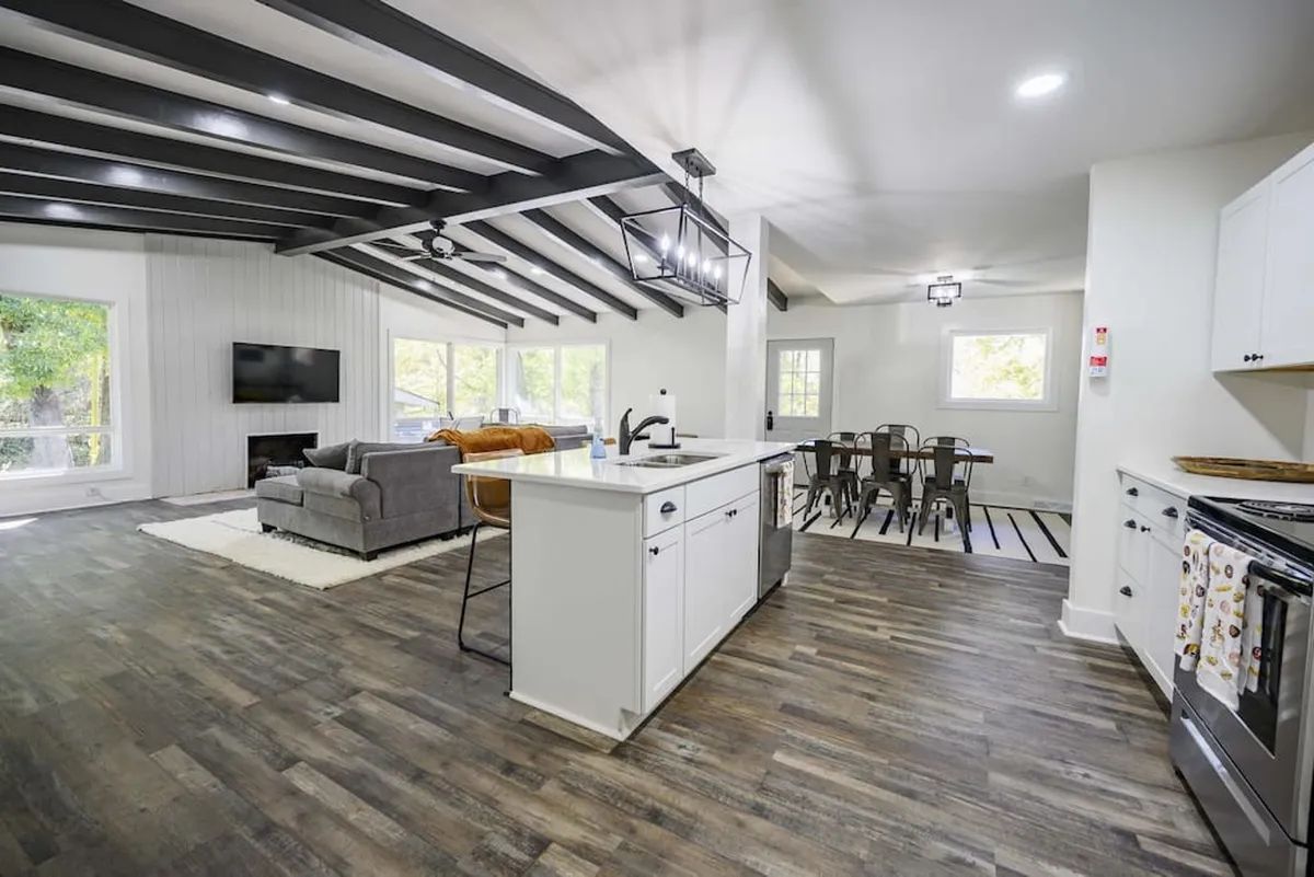This image captures a bright, open-concept living area where a kitchen island, comfortable sofa, dining table, and large windows are visible. Natural light floods the space, highlighting both the communal kitchen and lounge zones beneath exposed beams.