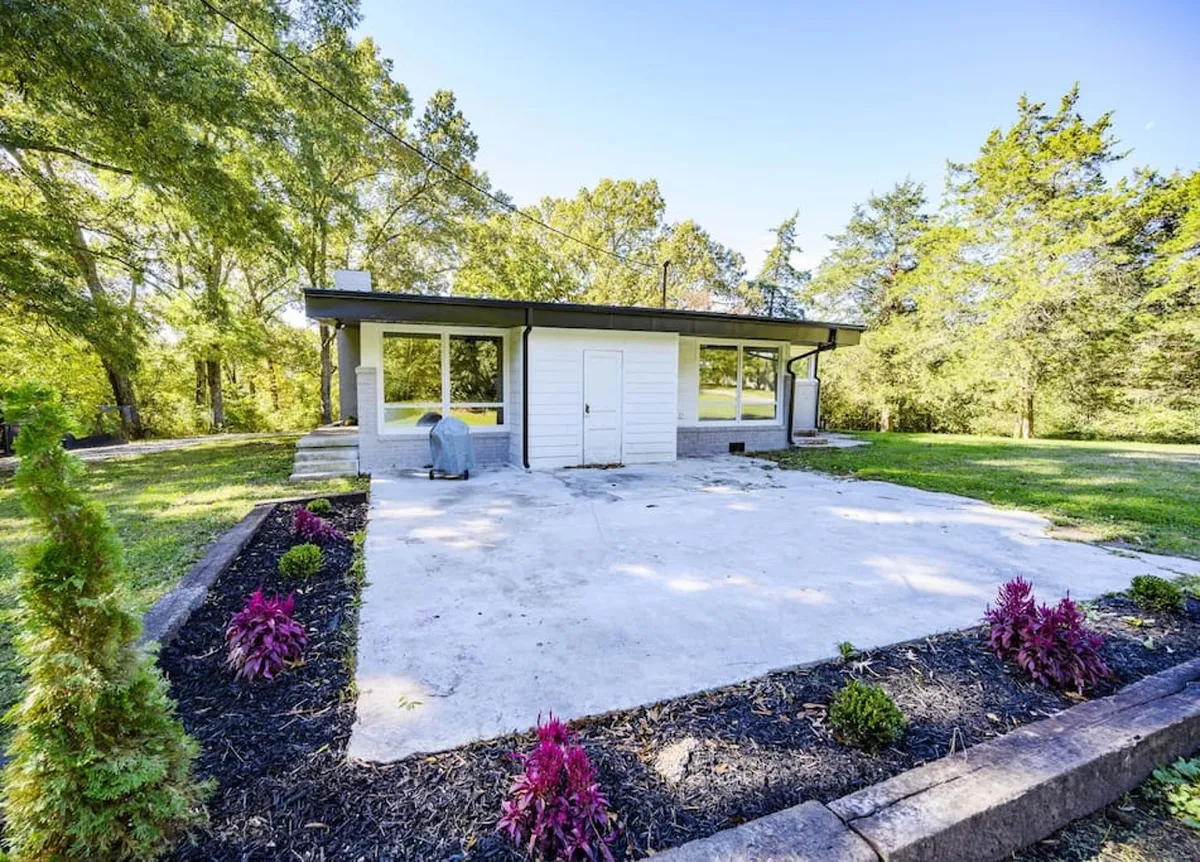 A manicured garden with a modern concrete patio set against the backdrop of the house's large windows and contemporary architecture. The open yard offers ample space for children to play or for guests to gather. The scene is bathed in natural light, enhancing the feeling of tranquility and spaciousness.